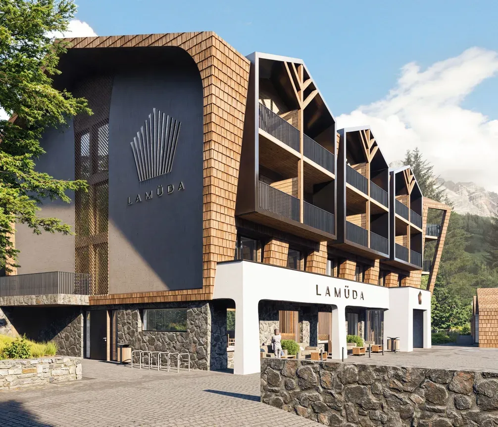 Contemporary multi-story hotel with wooden and dark panel exterior, balconies, and "Lamuďa" signage, set against a mountainous backdrop with trees and a bright sky.