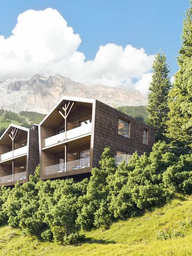 Modern wooden buildings with large balconies on a lush, green hillside, surrounded by trees and mountains in the background under a partly cloudy sky.