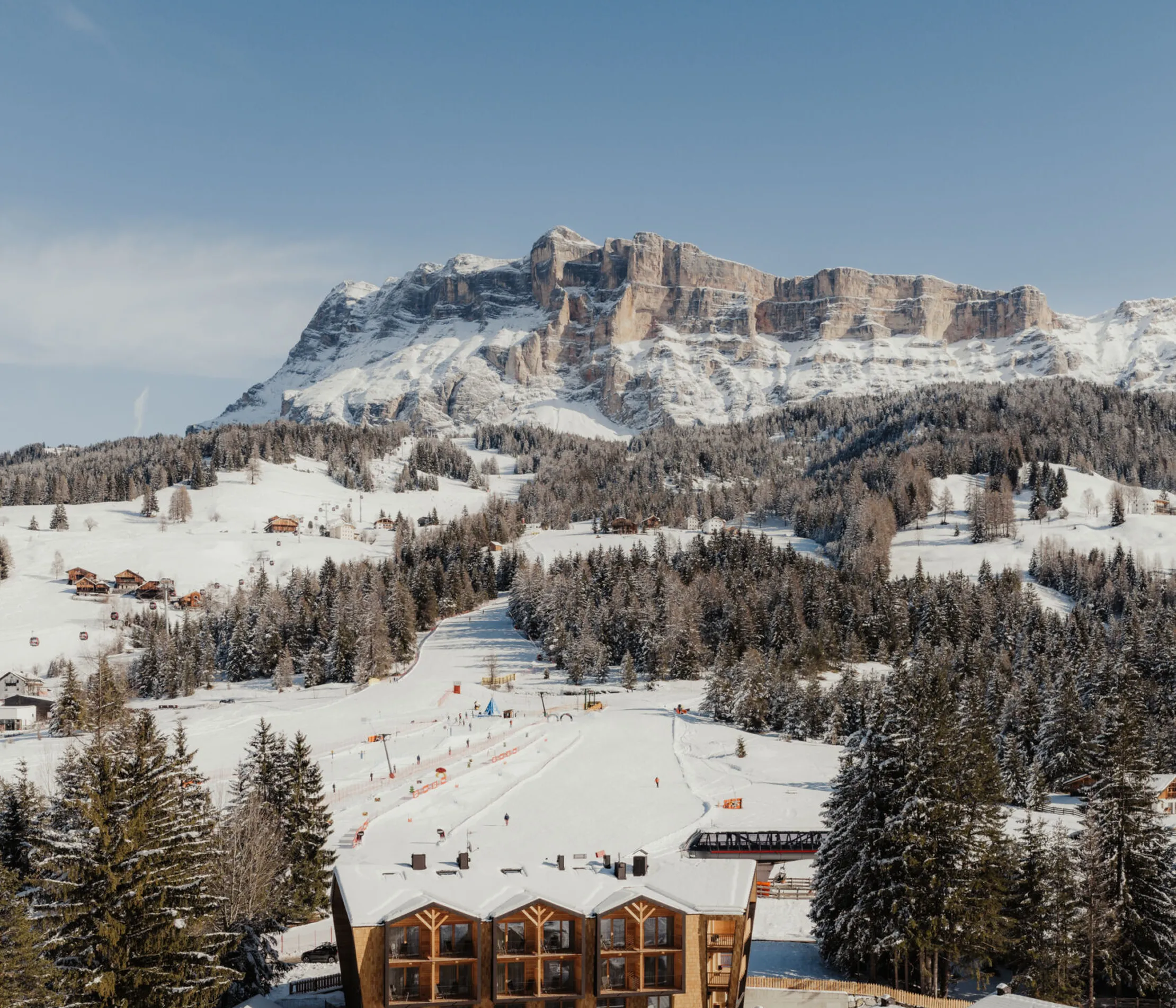 Verschneite Bergszene mit einem Skigebiet unten, rustikale Holzarchitektur umgeben von Bäumen. Ein majestätischer verschneiter Gipfel und blauer Himmel im Hintergrund.