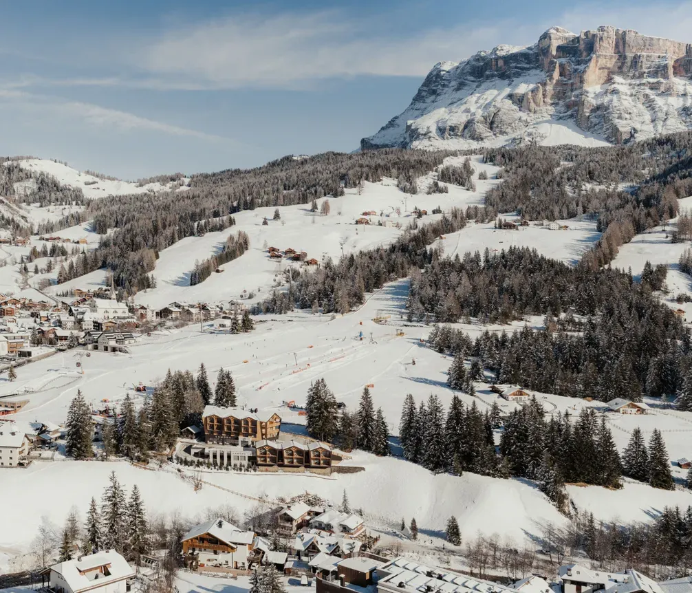Verschneites Alpendorf eingebettet in Kiefernwälder, mit Skiabfahrten, die sich durch die Landschaft schlängeln. Majestätischer Berggipfel im Hintergrund unter klarem blauem Himmel.
