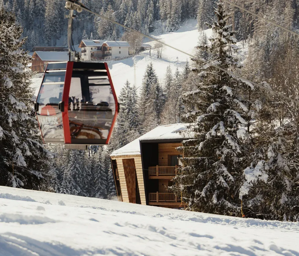Eine rote Seilbahn gleitet über eine verschneite Berglandschaft. Kiefern und Hütten liegen eingebettet im Schnee, unter einem klaren blauen Himmel, was winterliche Ruhe vermittelt.