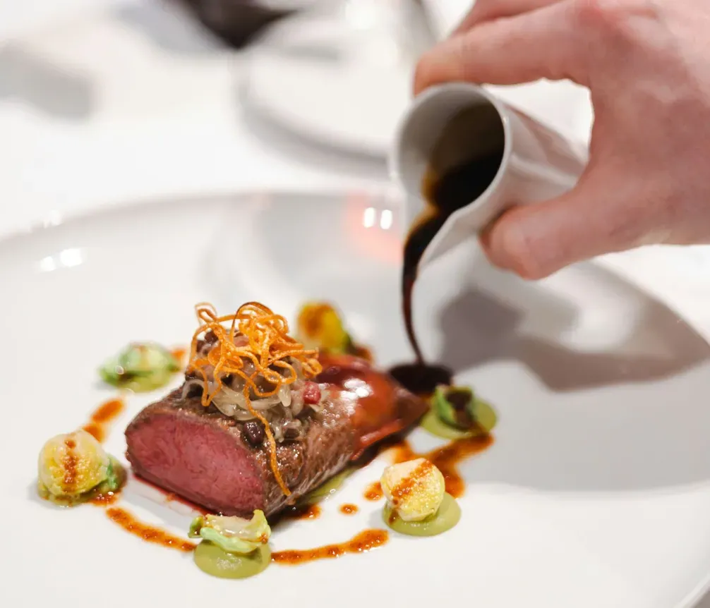A hand pours dark sauce over a cooked beef fillet garnished with crispy vegetables and dollops of green puree on a white plate, with glasses of red wine and a blurred background.