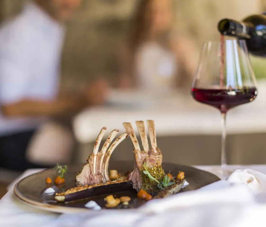 A gourmet dish featuring two lamb chops with exposed bones served on a dark plate, with a glass of red wine nearby, set on a white tablecloth at Hotel La Müda, Alta Badia.