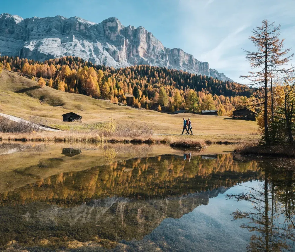 Autumn landscape with mountains, colorful forest, two hikers, and the reflection of the scenery in a lake.