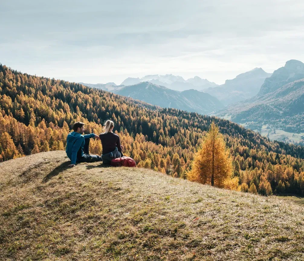 Two hikers sitting on a meadow with views of the mountains and autumn forests in the Dolomites, with rugged mountains in the background and a clear sky.
