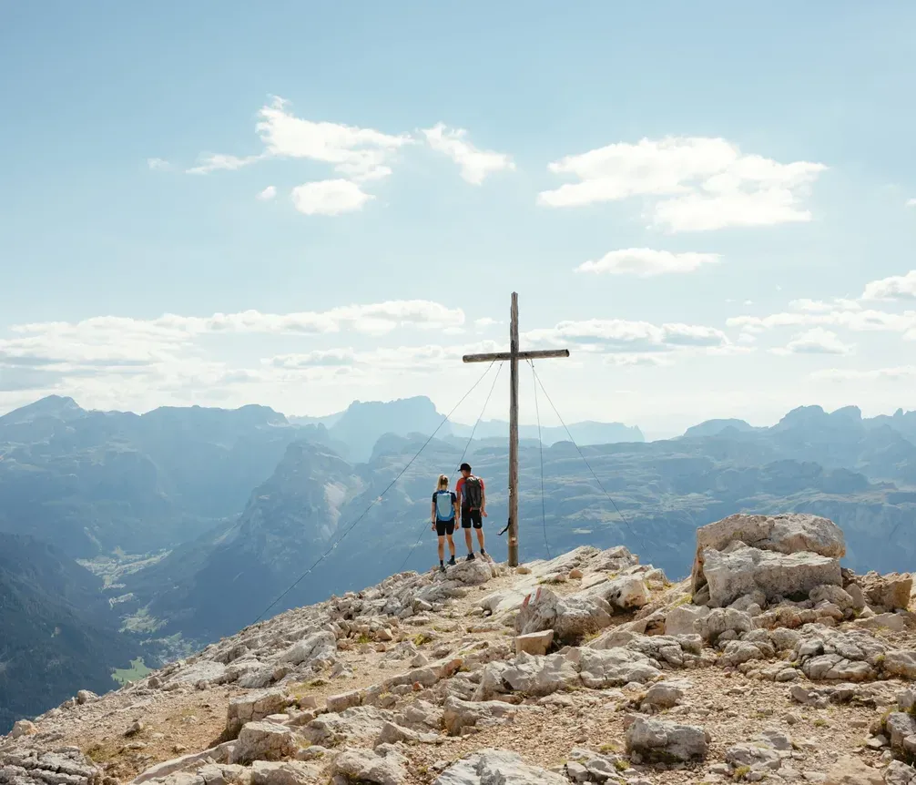 Two hikers enjoying the view of the Dolomites near a summit cross on a rocky peak, with mountains and clouds in the background.