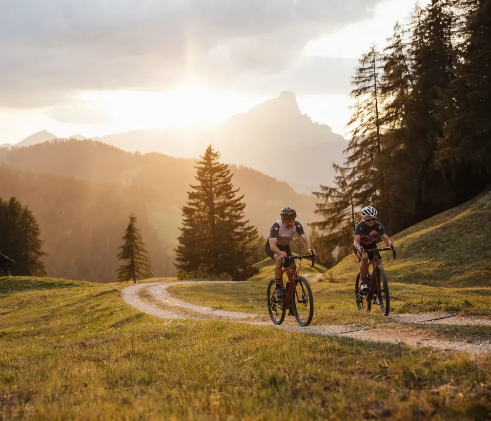 Two cyclists riding along a trail on a green plain, with trees and mountains in the background, at sunset in Alta Badia, Dolomites.