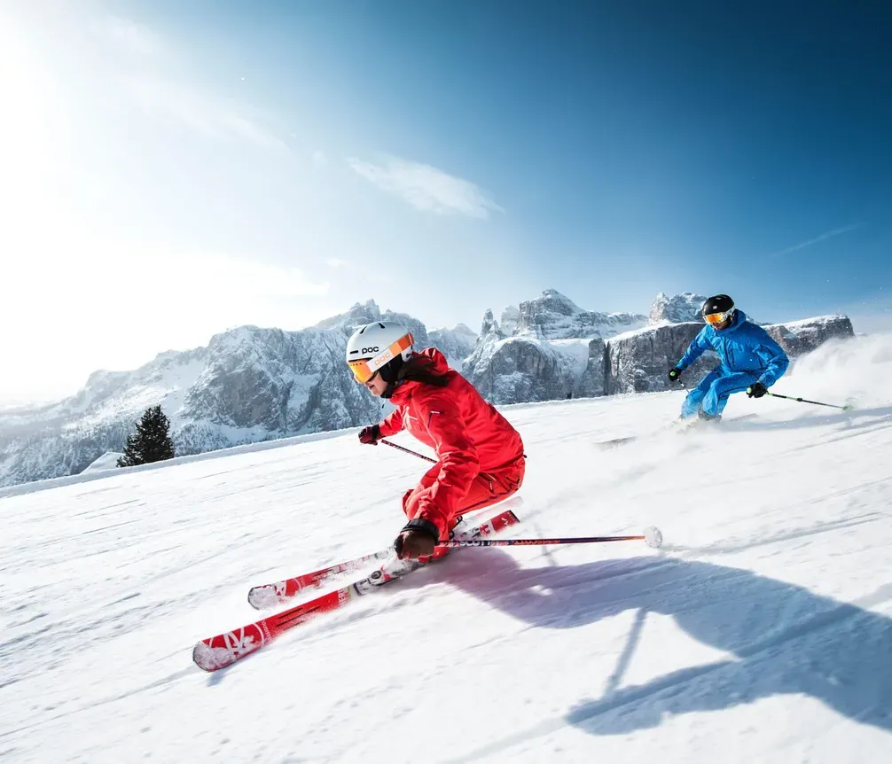 Two skiers on a snow slope, with mountains and forests in the background, under a blue sky at a ski resort in the Dolomites.