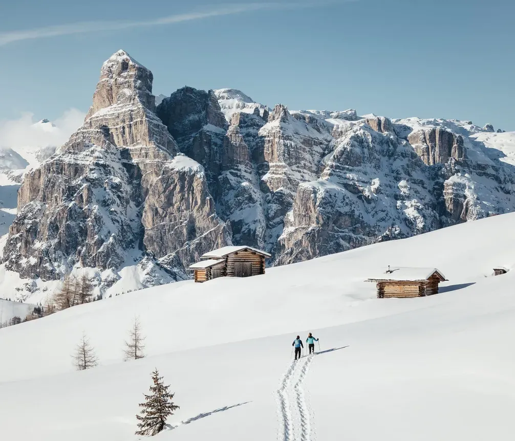 Two people walking in the snow with snowshoes, among simple wooden chalets in a snowy mountain landscape with majestic mountains in the background.