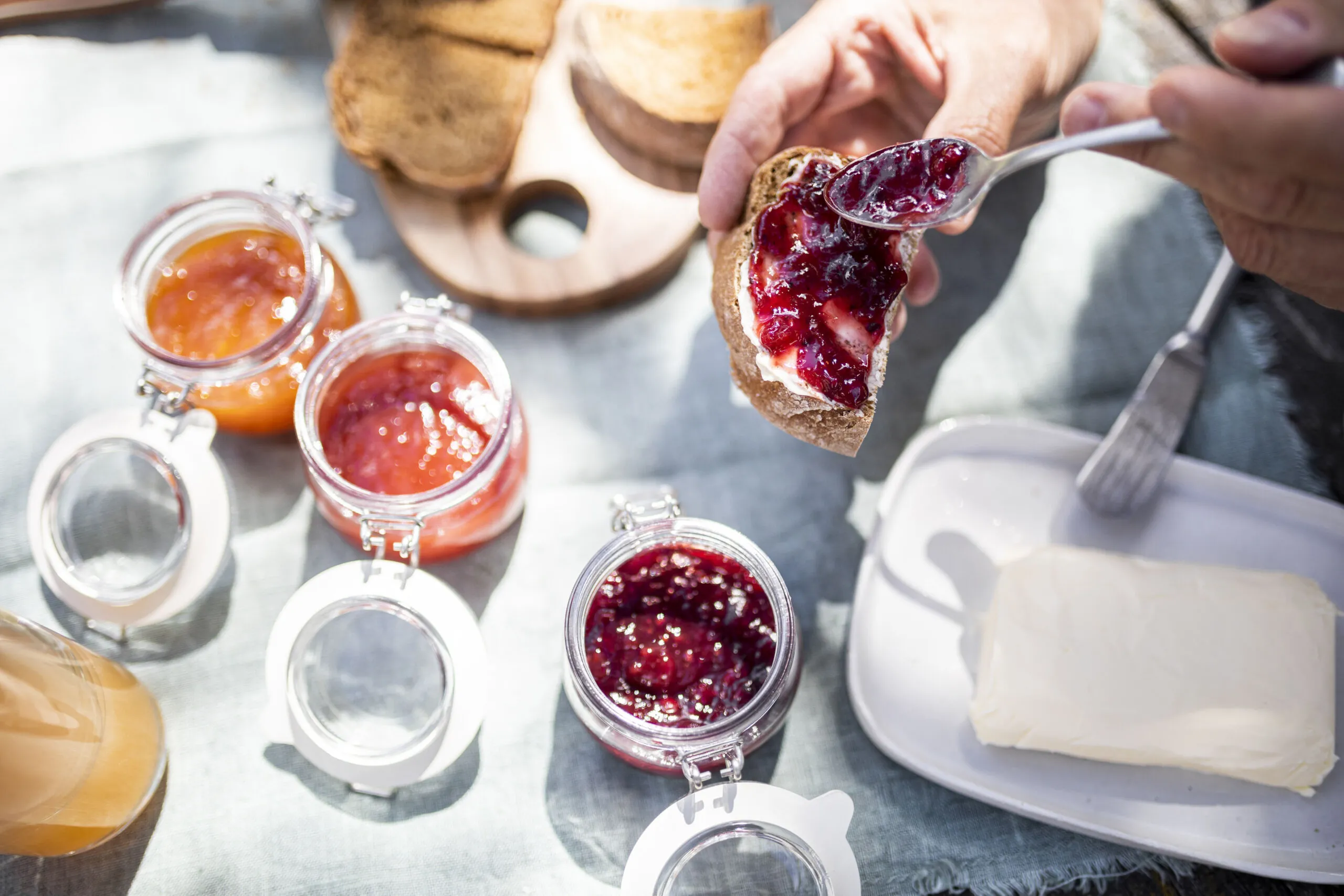 Pane con marmellata di frutta e barattoli di marmellata in un tavolo, con una mano che spalma marmellata su una fetta di pane.