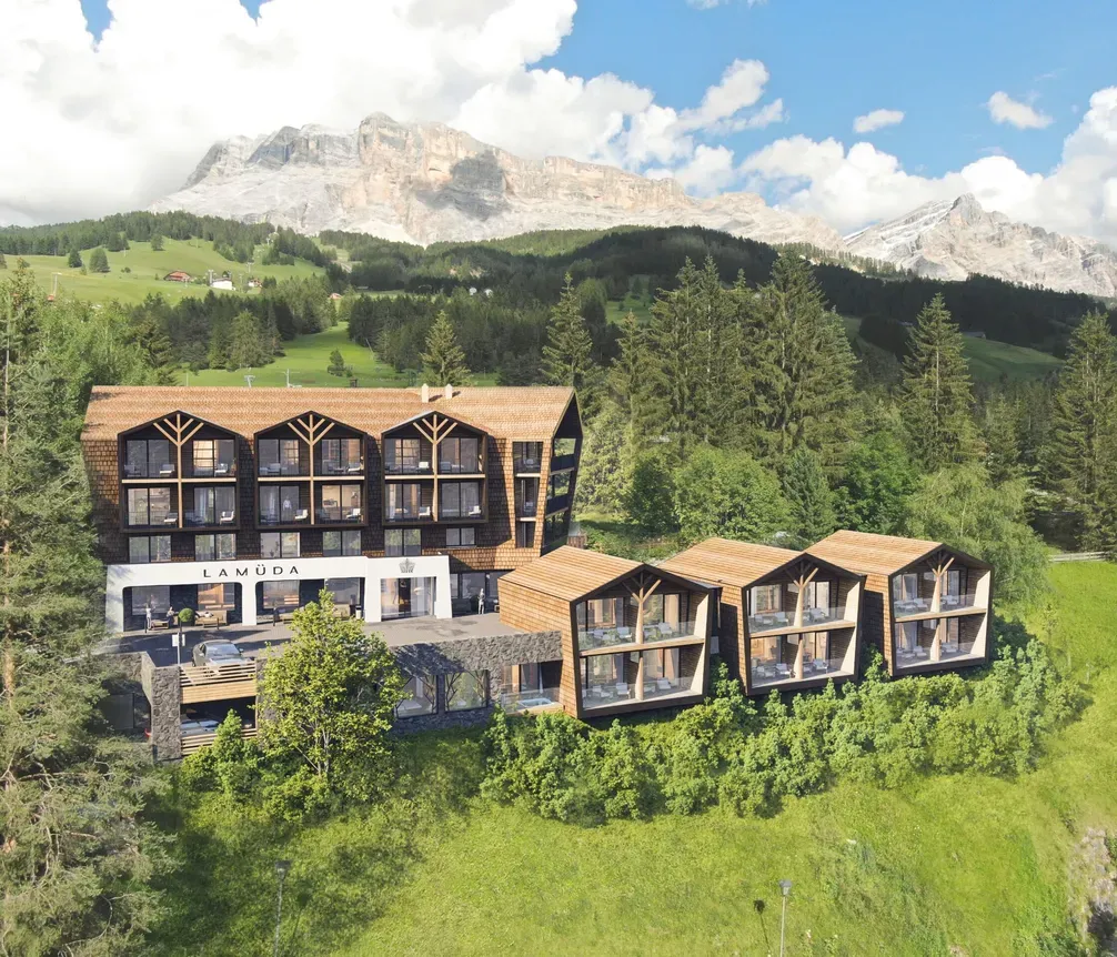 Modernes Hotel La Müda in Alta Badia, Dolomiten, auf grüner Wiese mit Bergblick, umgeben von Bäumen und Bergen im Hintergrund.