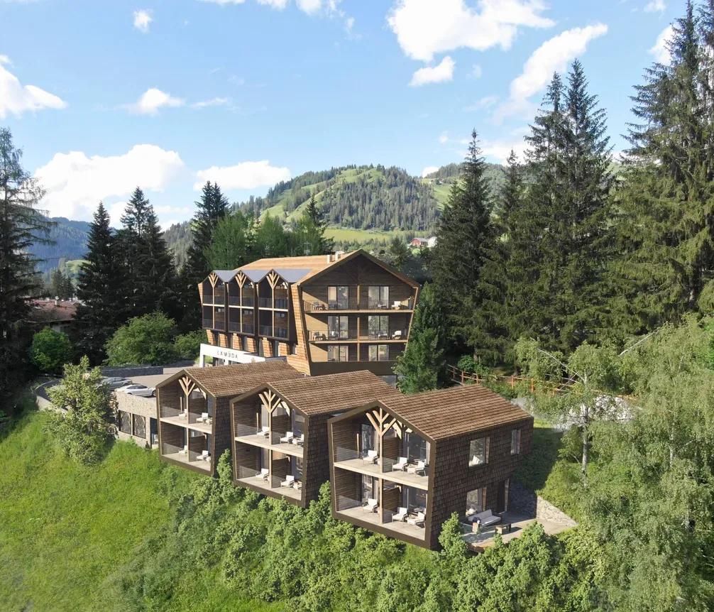 Moderne Hotelanlage in den Bergen von Alta Badia, Dolomiten, umgeben von grünen Wäldern und Hügeln bei sonnigem Himmel.