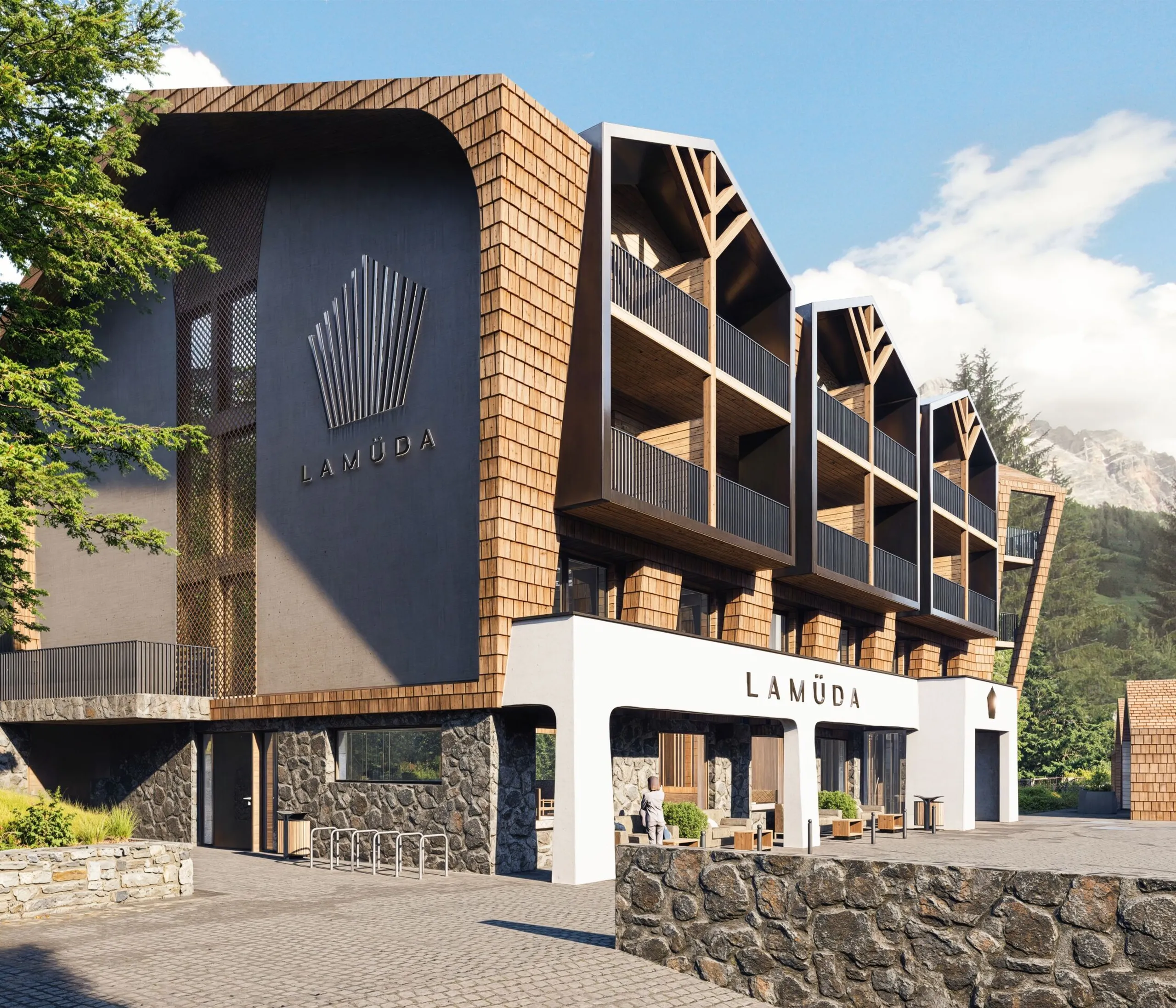 Edificio moderno con fasciate di legno e elementi di pietra, con logo di La Müda sulla parete, situato tra prati verdi e montagne nelle Dolomiti.