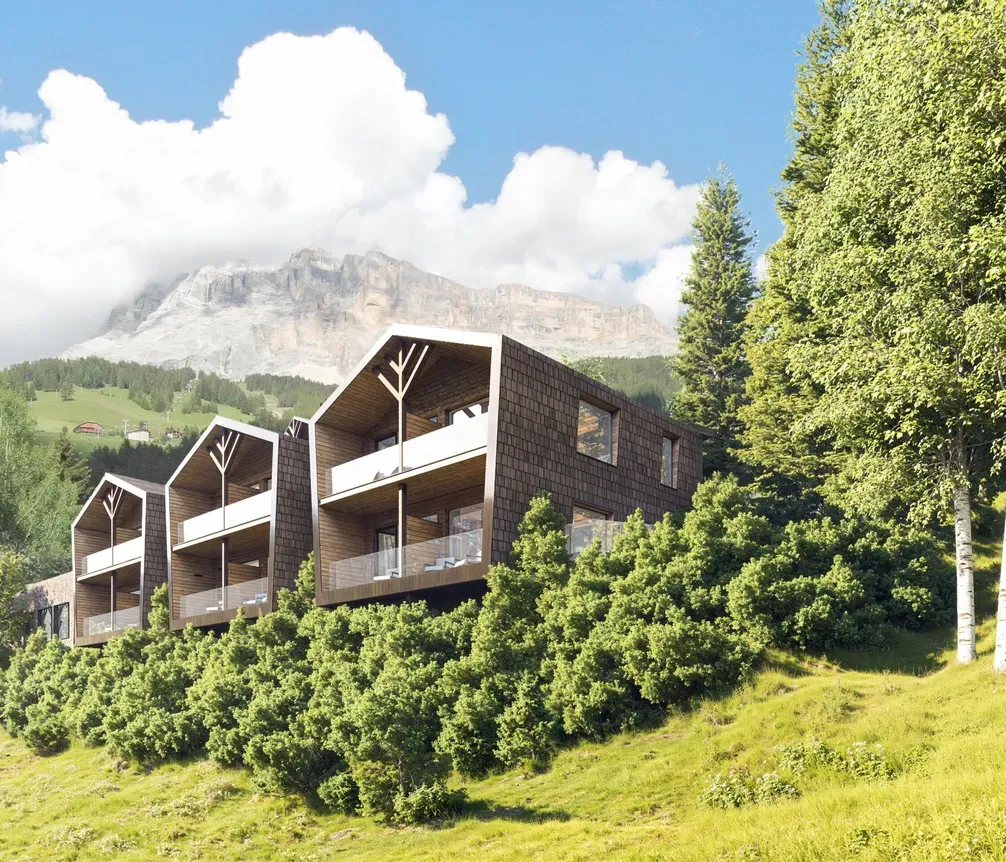 Moderne Gebäude mit Balkonen in grüner Hügellandschaft vor einer Bergkulisse, umgeben von Bäumen, in Alta Badia, Dolomiten.