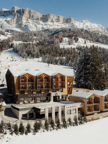 Snowy mountain resort with wooden architecture set against majestic peaks and pine forests. Clear blue sky completes a serene winter scene.