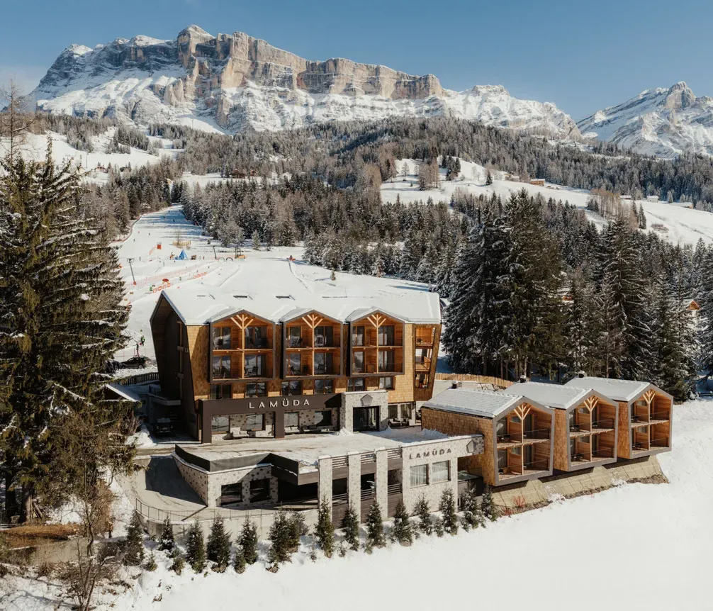 Snowy mountain resort with wooden architecture set against majestic peaks and pine forests. Clear blue sky completes a serene winter scene.