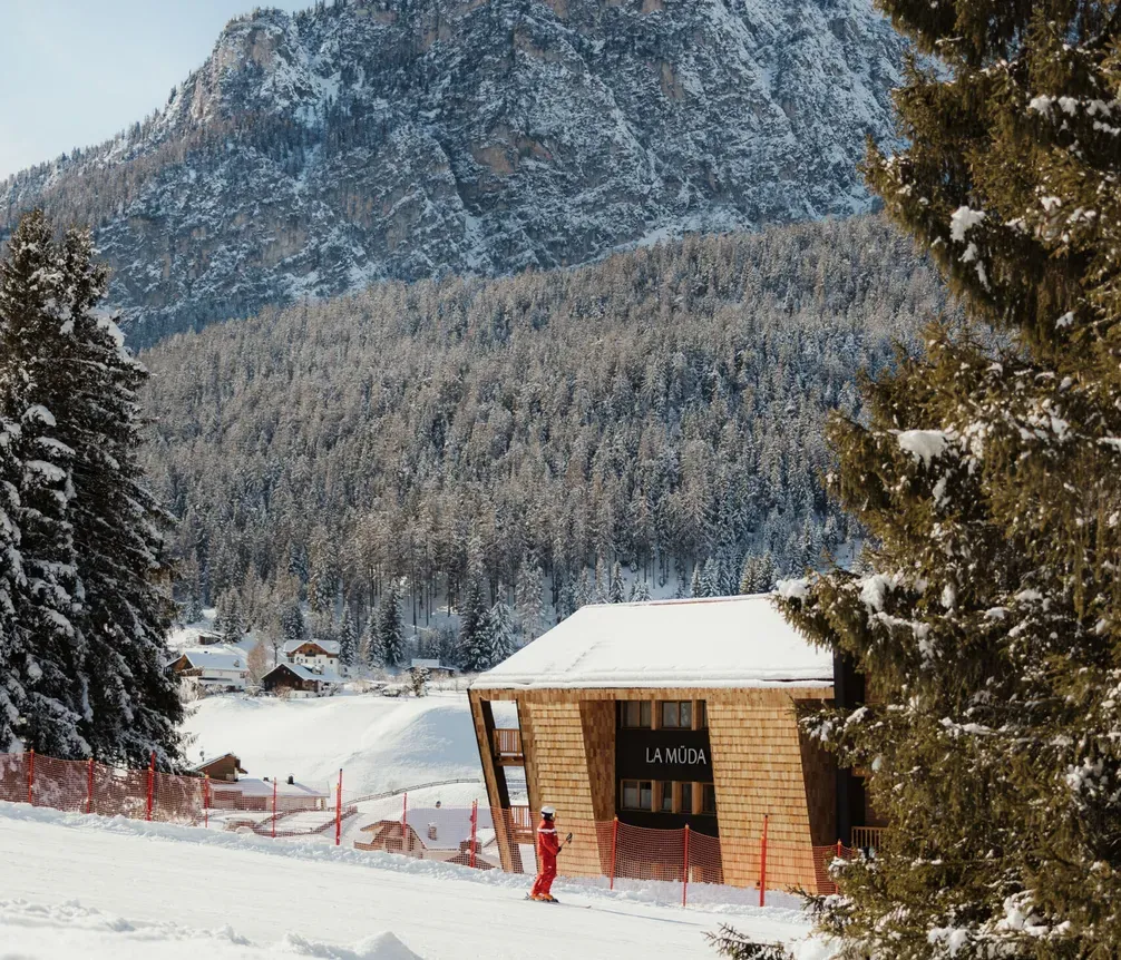 Snowy landscape with a wooden building marked "La Mida" next to pine trees. A person in red snow gear stands nearby. Majestic mountains in the background.