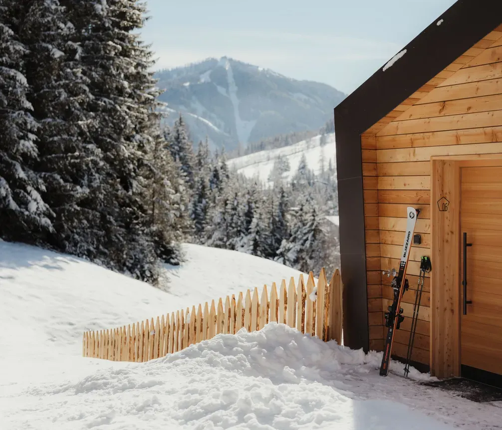 A wooden cabin in a snowy landscape, pine trees, and mountains in the background. Skis lean against the cabin, conveying a serene, winter getaway mood.