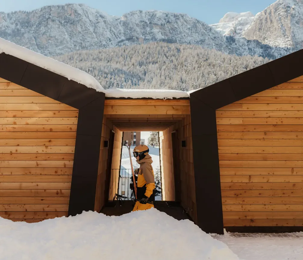 A skier in yellow pants and a helmet walks through a snow-covered wooden structure, with majestic mountains in the background under a clear blue sky.
