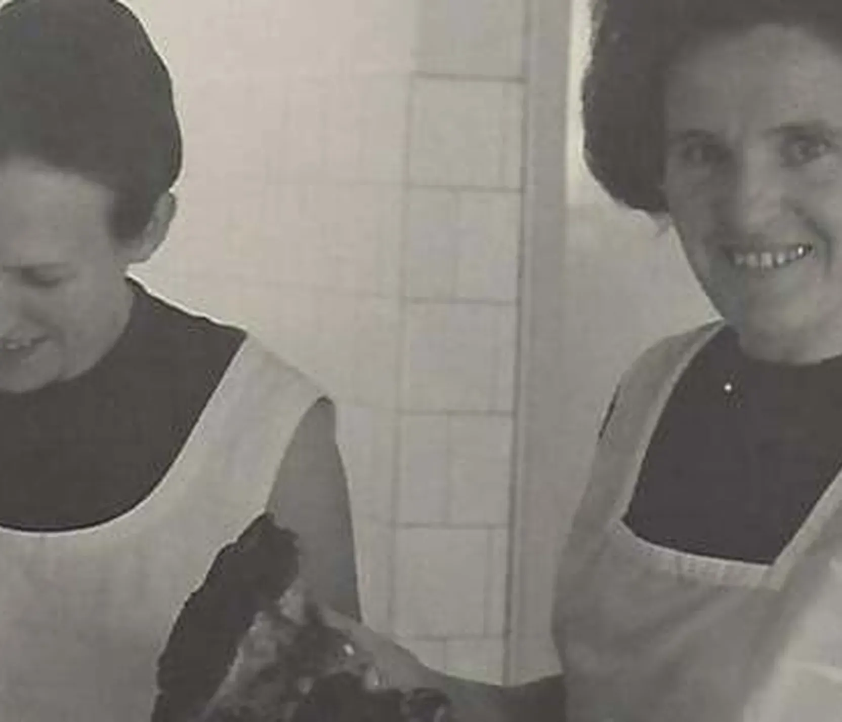 Zwei Frauen in Küchenschürzen bereiten gemeinsam Essen zu, lachen und wirken fröhlich in einer Küchenumgebung.