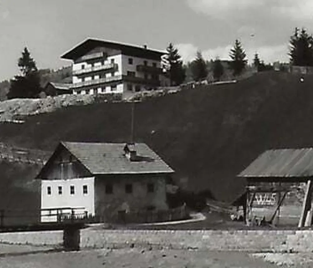 Dorf mit traditionellen Holzhütten und modernem Hotel im Hintergrund in Alta Badia, Dolomiten, unter bewölktem Himmel.