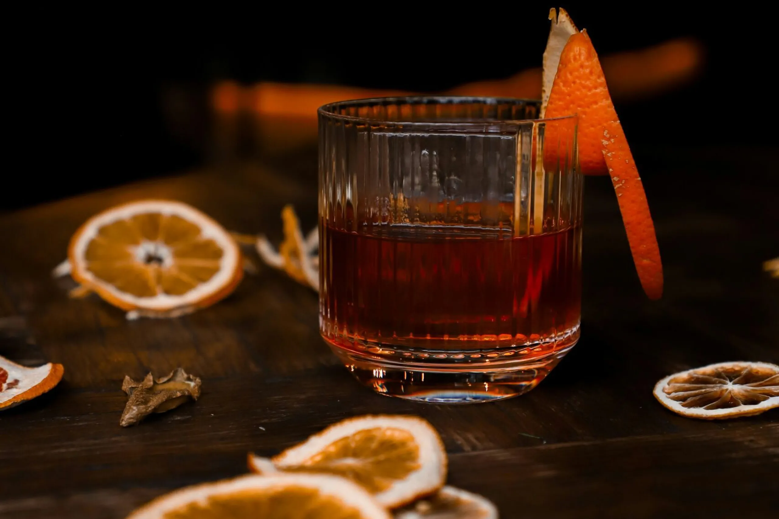 Un bicchiere di cocktail rosso con una fetta di agrume decorativa, circondato da fette di arancia secche e scorze di agrume su un tavolo di legno scuro.