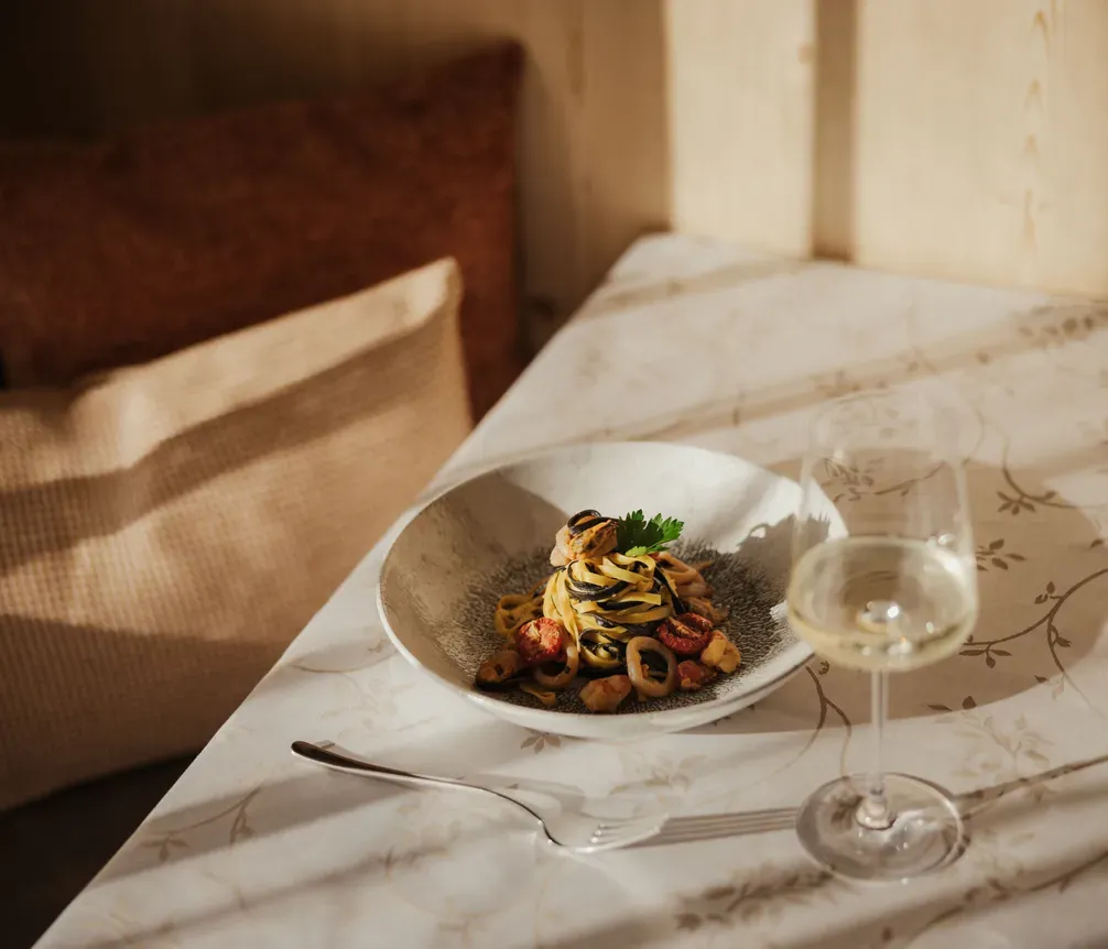A bowl of pasta with cherry tomatoes and garnish is on a sunlit table with a fork and a glass of white wine, evoking a cozy, elegant ambiance.