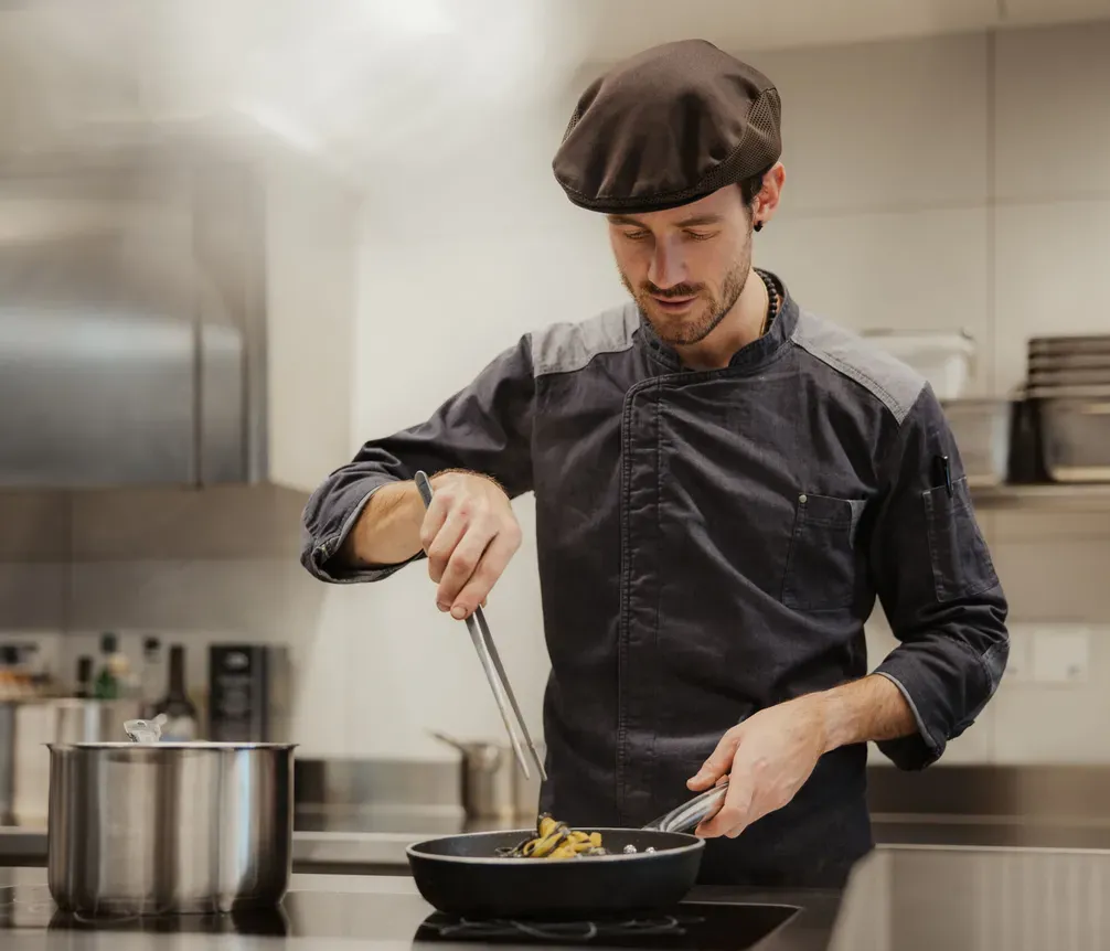 Chef in a dark uniform and cap cooks with focus in a sleek kitchen. He uses tongs over a pan, exuding calm and professionalism. Steam rises nearby.