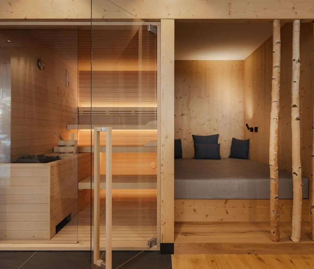 Modern spa room with a glass-enclosed sauna on the left, featuring warm wood tones and soft lighting. Cozy relaxation nook with cushions on the right.