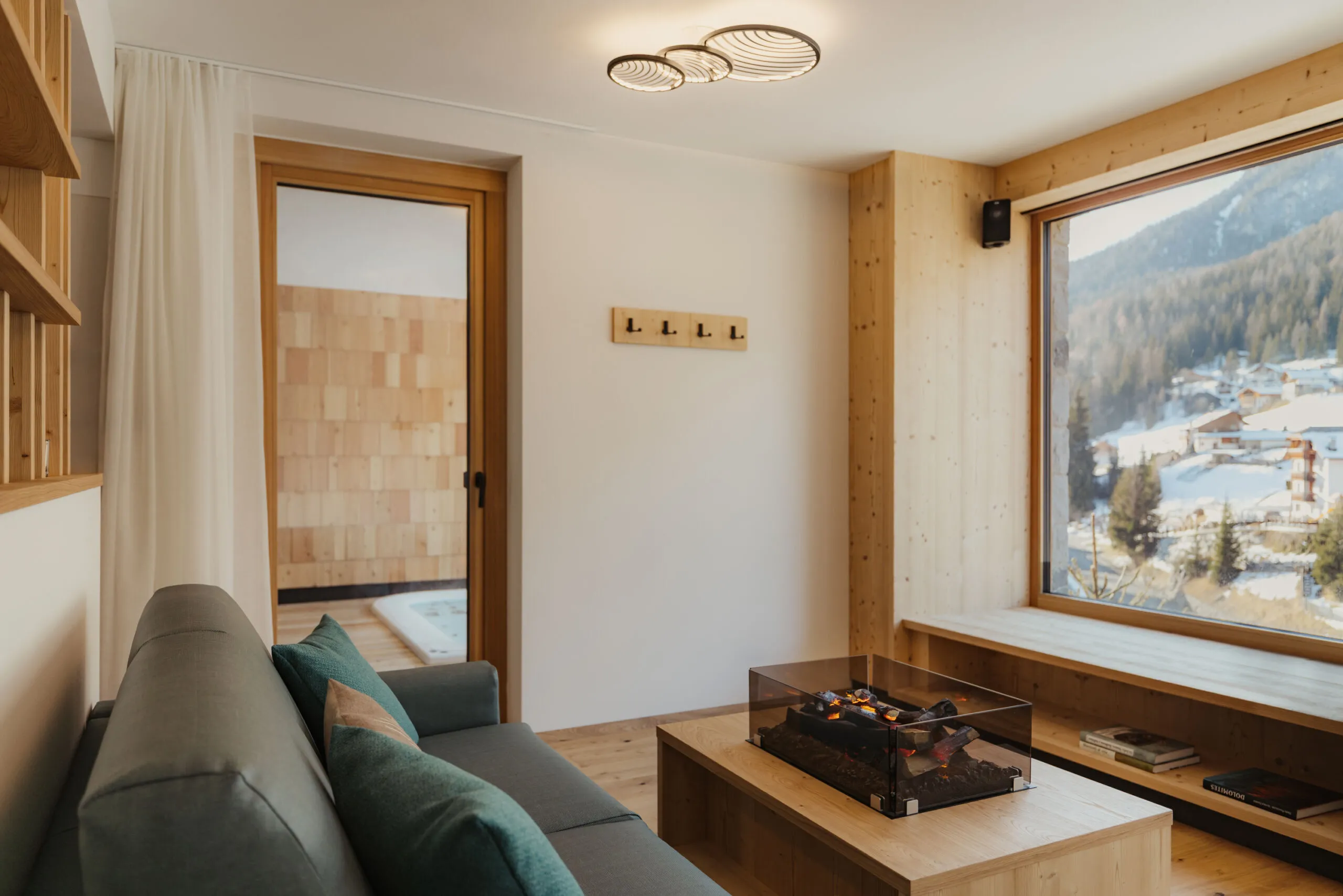 Cozy living room with large window overlooking snowy mountains. Modern decor with a green sofa, glass fireplace, and light wood accents. Peaceful ambiance.