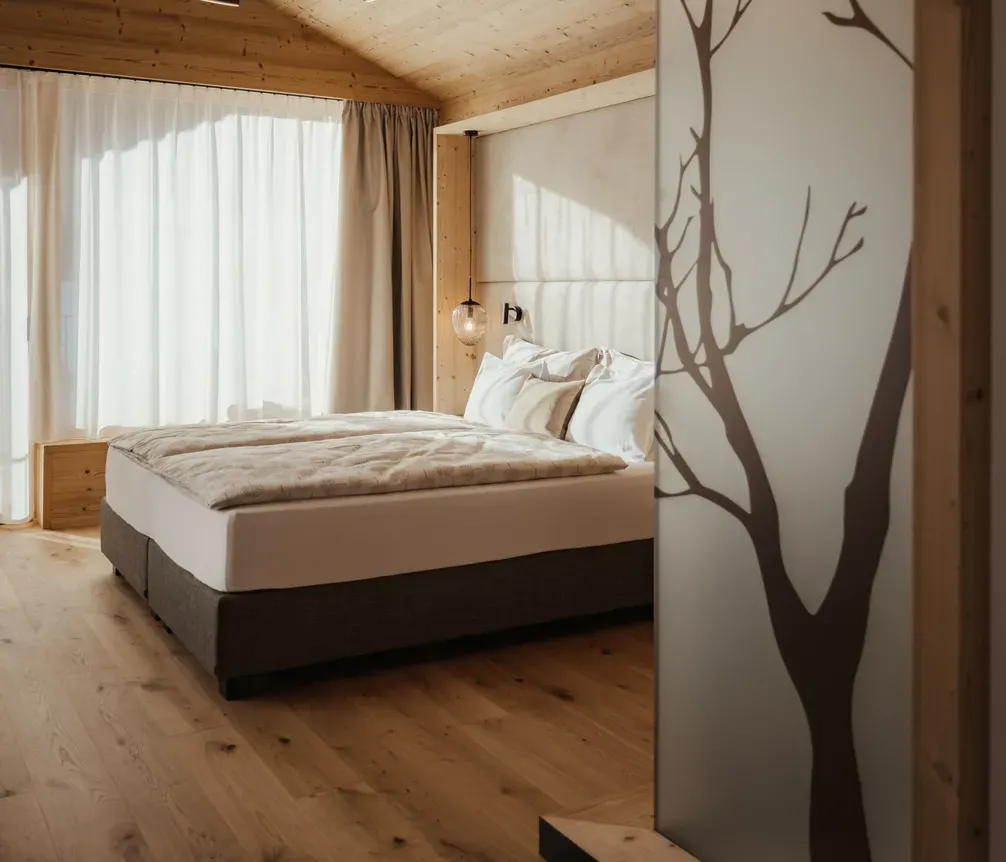 A cozy bedroom with a wooden ceiling and floor, soft lighting, and a frosted glass partition featuring a tree design. Natural light filters through curtains.
