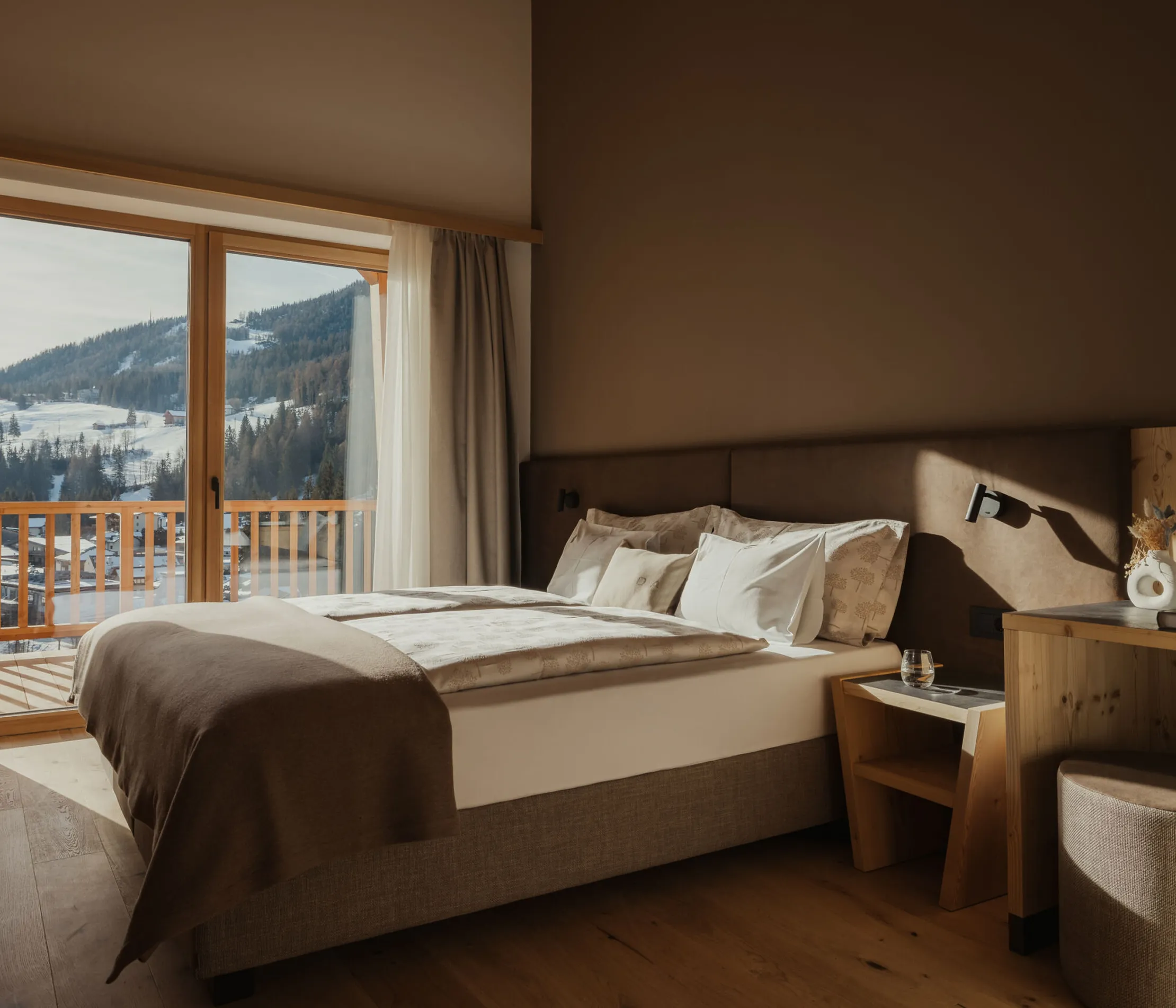 Cozy bedroom with a large window showcasing a scenic mountain view. The room features a neatly made bed, wooden furniture, and soft lighting, creating a warm, inviting atmosphere.