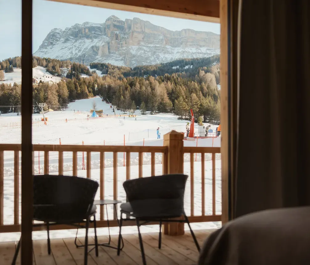 View from a cozy room showcasing a snowy ski resort scene with skiers, framed by a wooden balcony railing and distant snowy mountains.