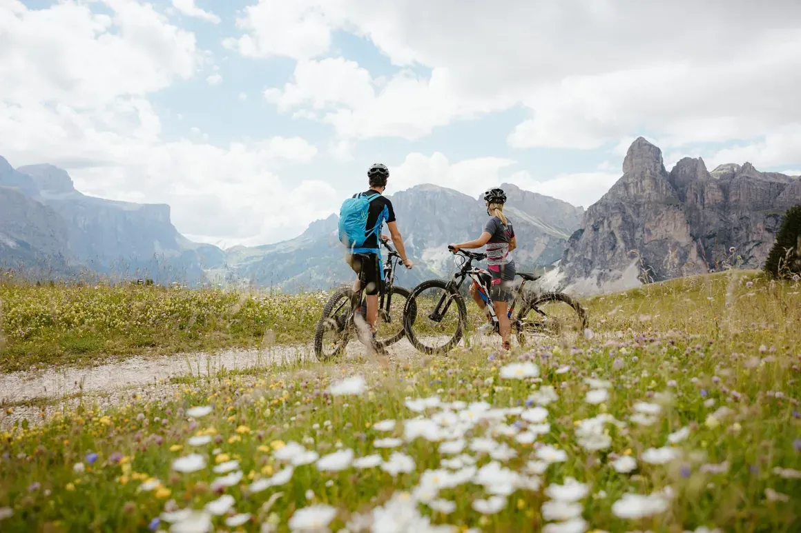 Due escursionisti in mountain bike su un sentiero tra prati fioriti con montagne imponenti sullo sfondo in Alta Badia, Dolomiti.