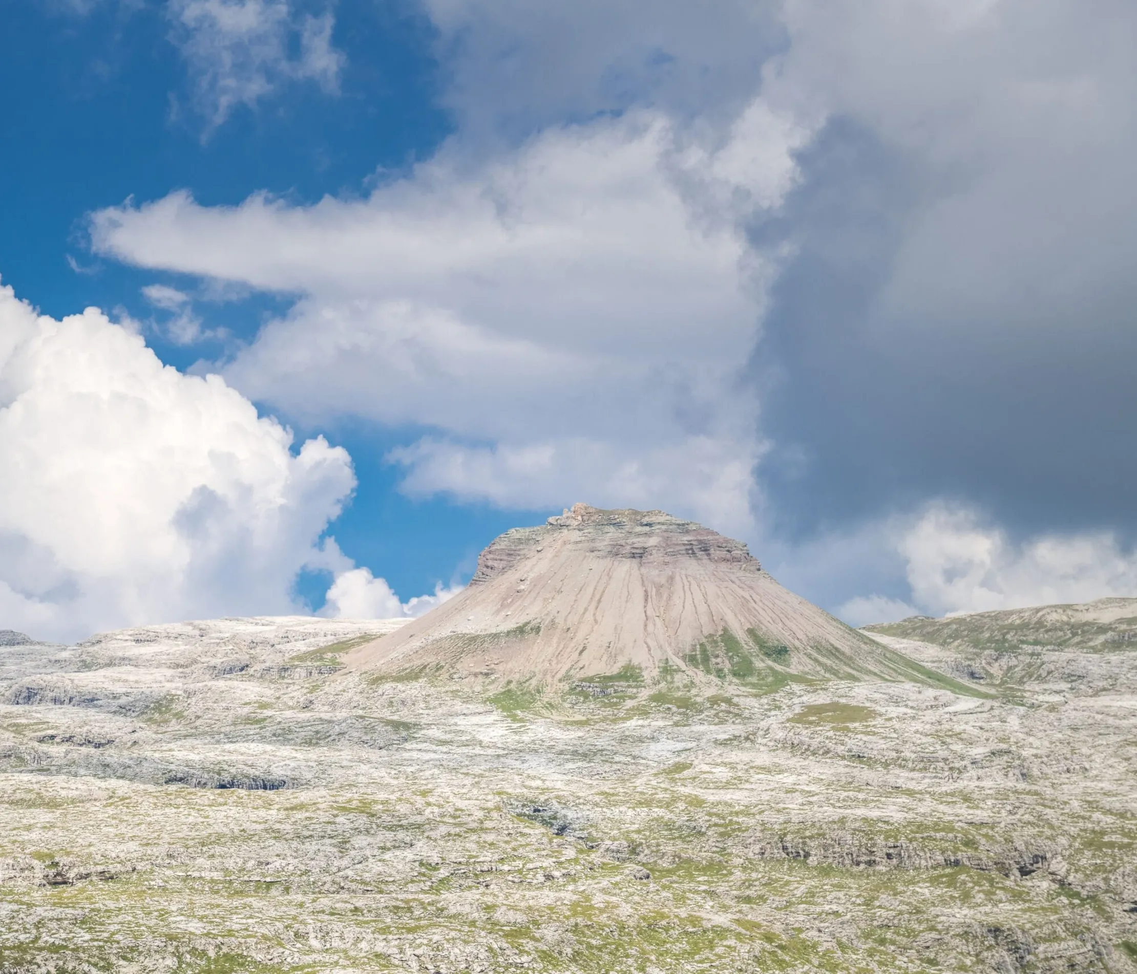 A rocky mountain formation with slopes covered in grass and rock, under a partly cloudy sky in the Dolomites, Alta Badia region.