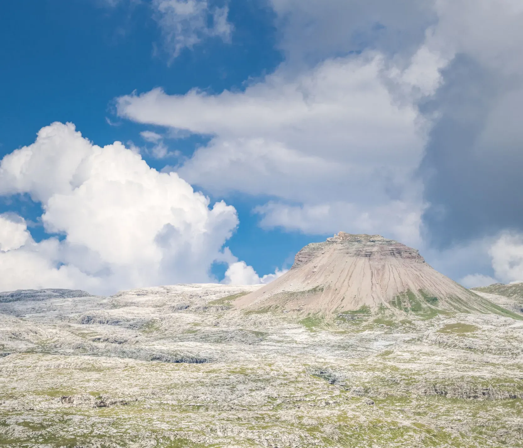 A rocky mountain formation with slopes covered in grass and rock, under a partly cloudy sky in the Dolomites, Alta Badia region.