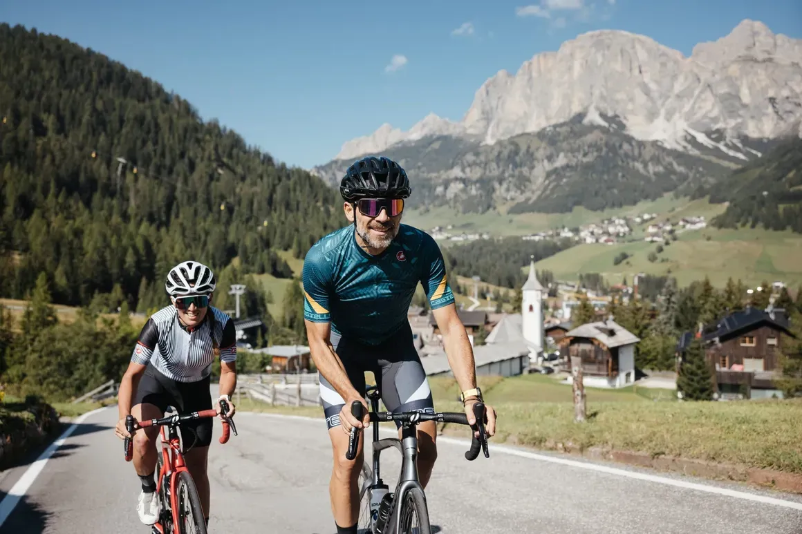 Due ciclisti pedalano su una strada tra montagne verdi e un paesino con chiesa, con imponenti rocce calcaree sullo sfondo nelle Dolomiti.