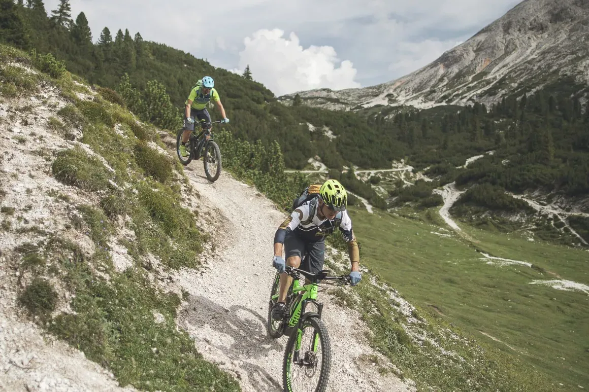 Due mountain bikers descend a rocky trail in the Dolomites, with green vegetation, a mountain in the background, and a partly cloudy sky above.