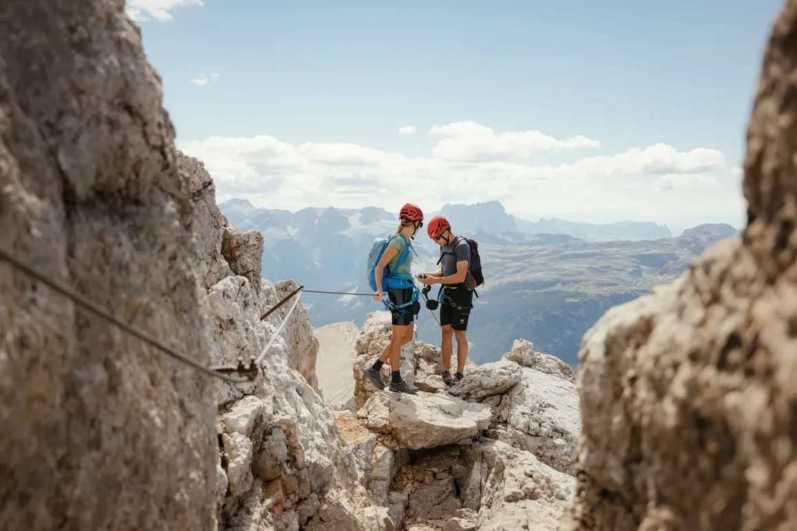 Due escursionisti con caschi rossi si trovano su una scalata tra le rocce con una vista sulle montagne e il cielo azzurro sullo sfondo.