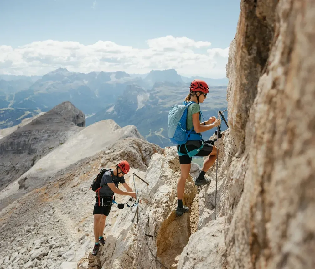 Zwei Kletterer mit Helmen und Ausrüstung steigen in den Dolomiten auf felsigem Gelände empor, Blick auf Berge und Wolken im Hintergrund.