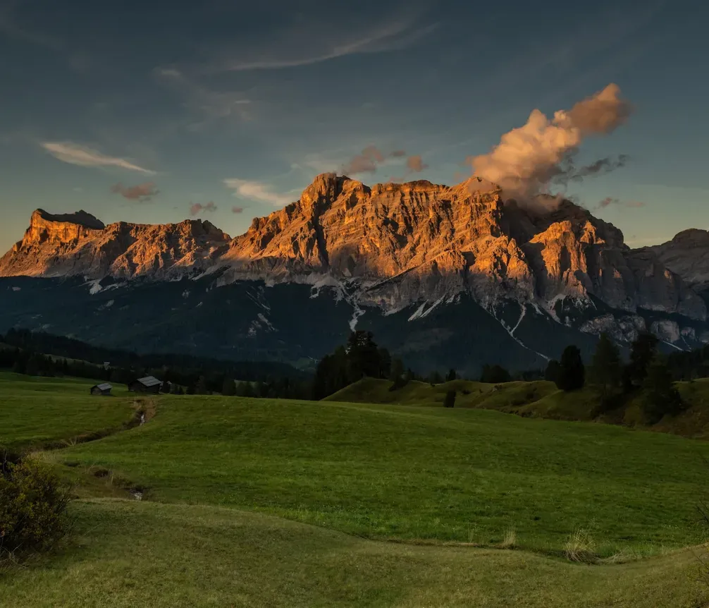 Majestätische Bergkulisse in Sonnenlicht, grüne Wiesen im Vordergrund, einzelne Hütten und kleine Bäche sichtbar, Himmel mit vereinzelt Wolken.