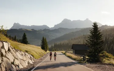 Due ciclisti pedalano sulla strada di montagna circondata da alberi e montagne nel paesaggio delle Dolomiti in Alto Adige, con cielo sereno e luce del tramonto.