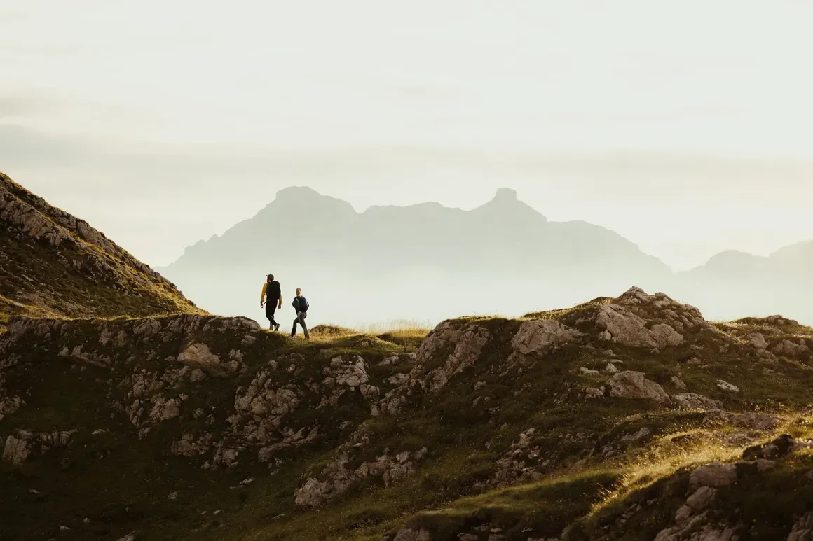 Due escursionisti camminano su un sentiero tra le montagne delle Dolomiti, con un paesaggio montano sfocato sullo sfondo in una luce soffusa.