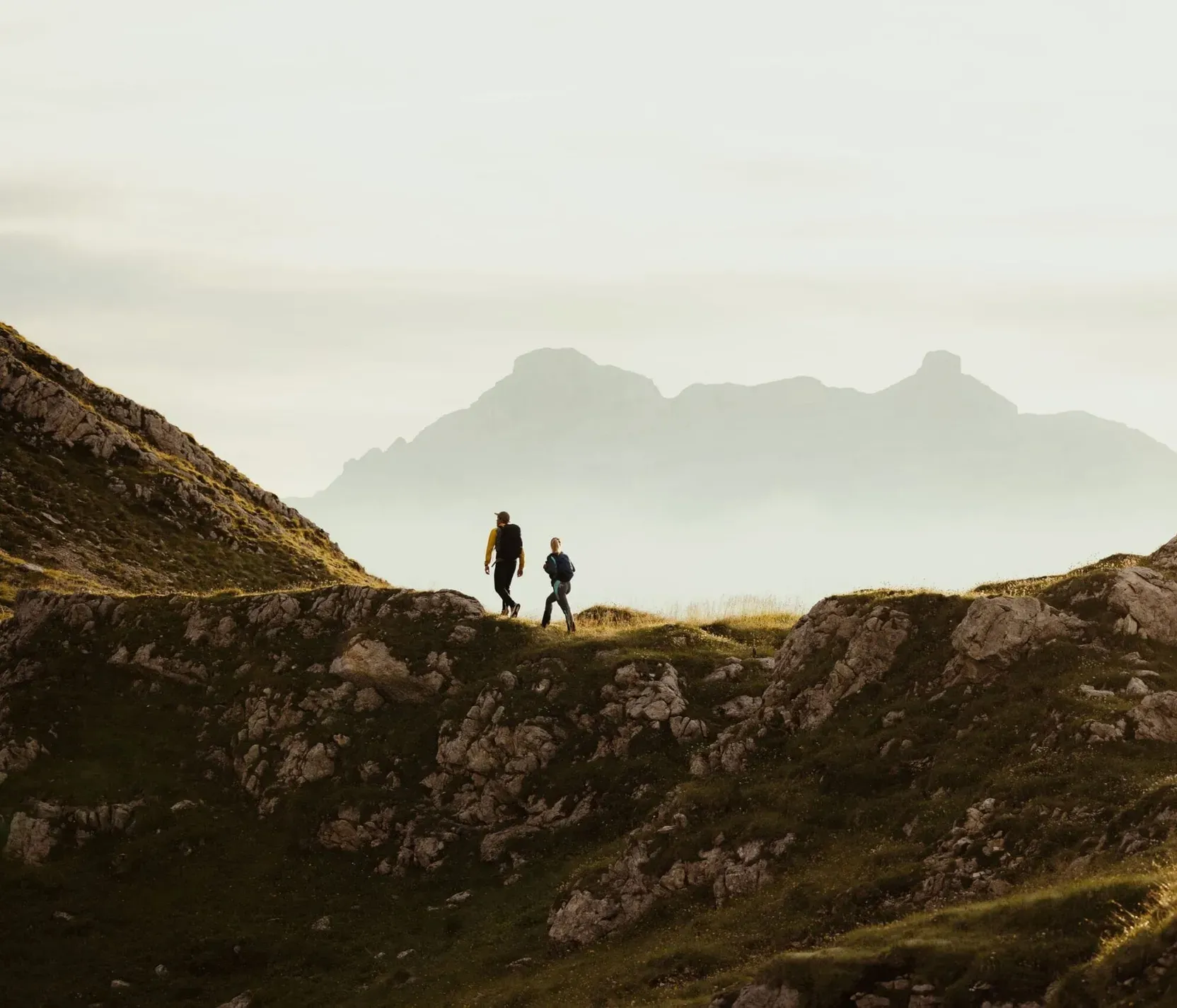 Two hikers walking on a trail among the Dolomites mountains, with a blurred mountain landscape in the background in soft light.