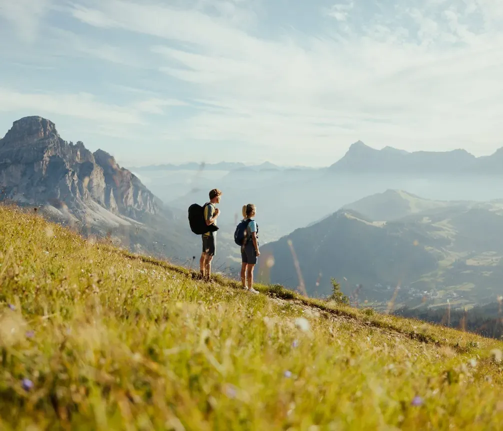 Zwei Wanderer mit Rucksack auf einer grünen Bergwiese mit Blick auf die Dolomiten im Hintergrund bei Sonnenaufgang.