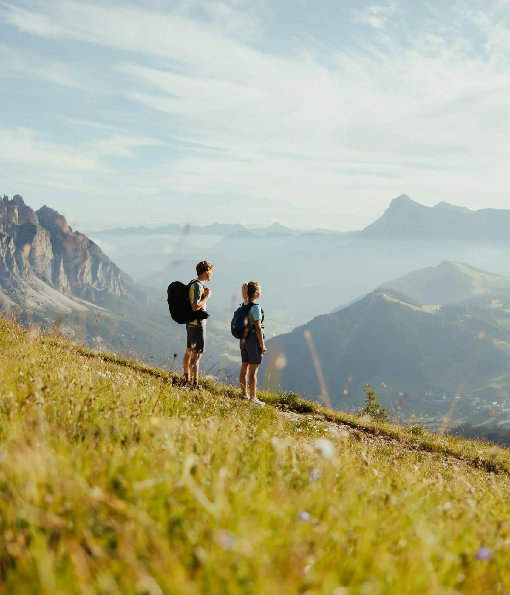 Zwei Wanderer mit Rucksack auf einer grünen Bergwiese mit Blick auf die Dolomiten im Hintergrund bei Sonnenaufgang.