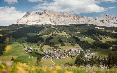 Paesaggio di montagne con villaggio di case tra verdi colline, con alcune case e alberi, sotto un cielo con nuvole.