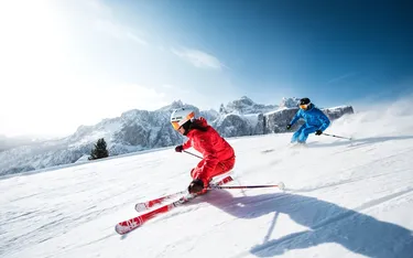 Zwei Skifahrer in bunten Anzügen fahren mit hoher Geschwindigkeit durch den Schnee vor einer Bergkulisse in den Dolomiten, Sonne und blauer Himmel im Hintergrund.
