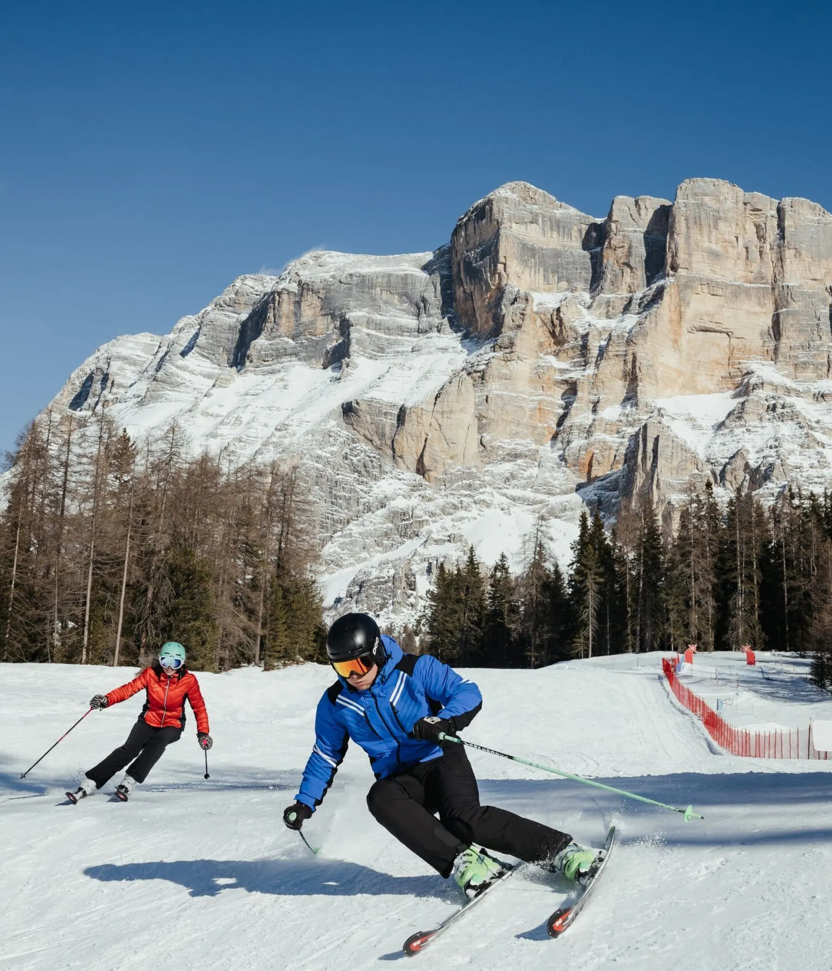 Due sciatori in pista sulla neve, con montagne e boschi sullo sfondo, sotto un cielo azzurro in una località sciistica delle Dolomiti.