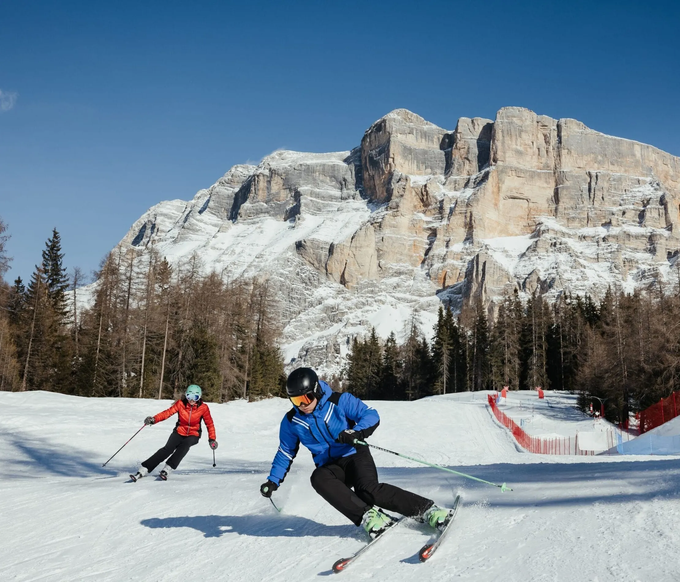 Two skiers on a snow slope, with mountains and forests in the background, under a blue sky at a ski resort in the Dolomites.